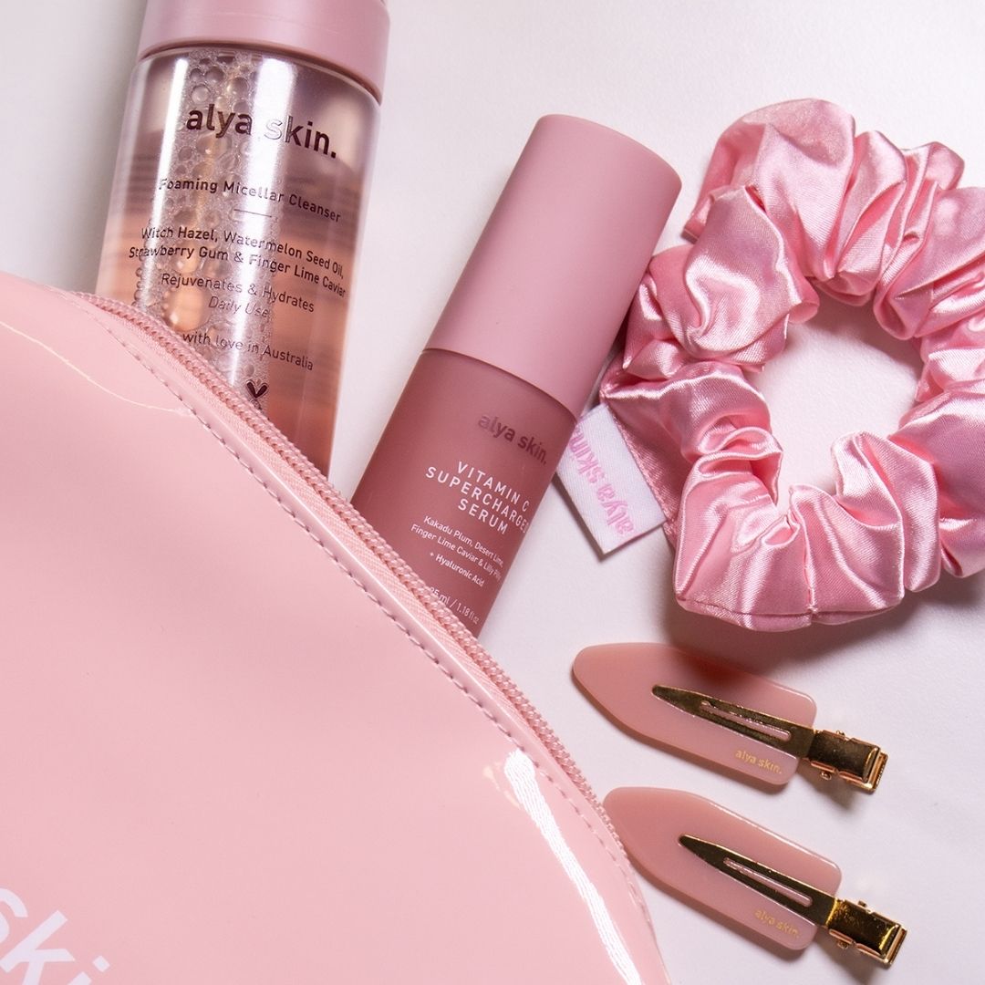 A skincare kit including a Foaming Micellar Cleanser and Vitamin C Supercharged Serum from Alya Skin, alongside a pink satin scrunchie and hair clips, presented on a white background.